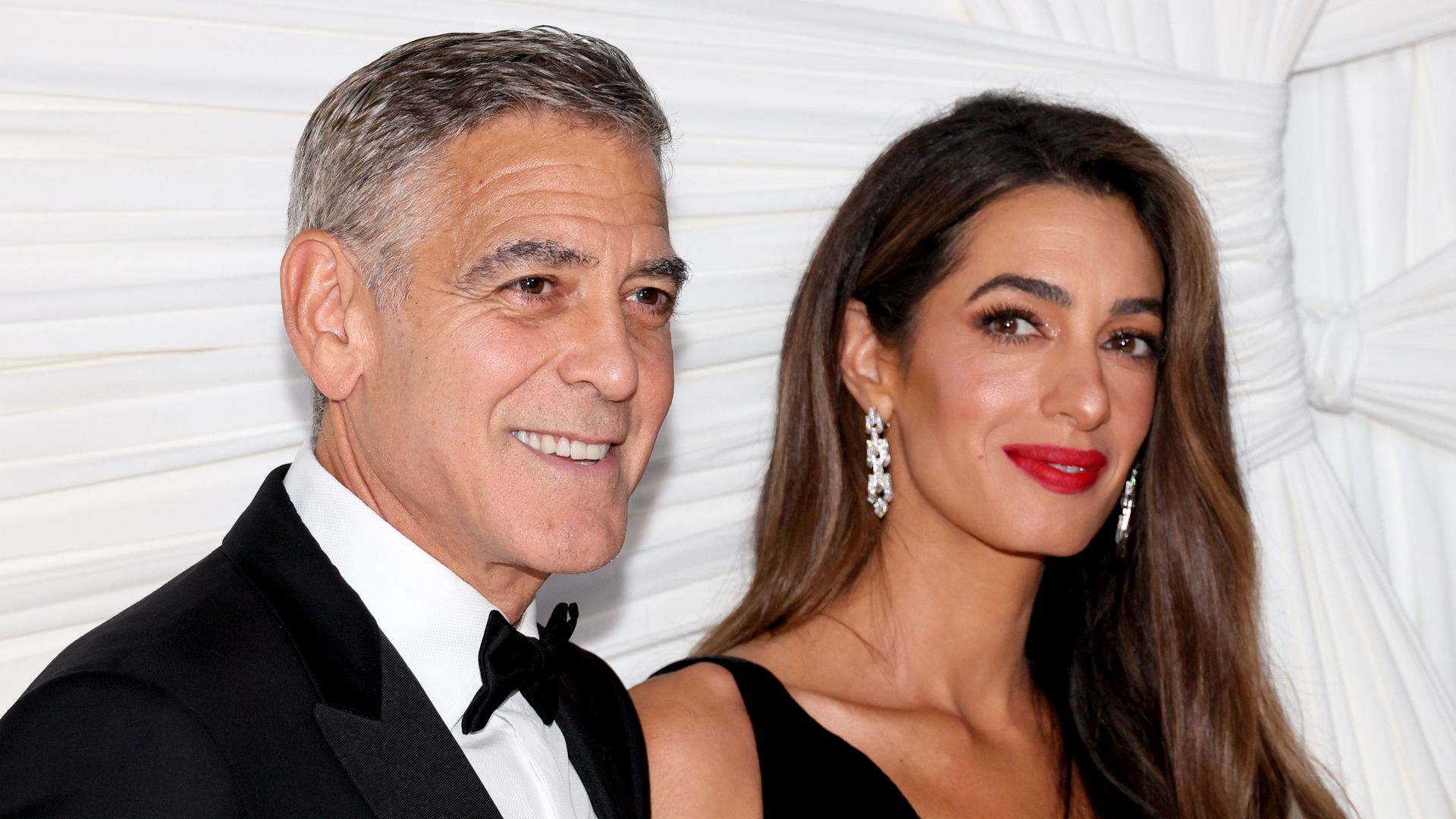 A man in a black tuxedo and bow tie stands next to a woman in a sleeveless black dress with long brown hair and red lipstick. Both are smiling and posing in front of a white draped background.