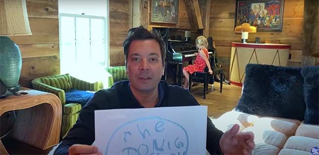 jimmy looks at the camera and holds up a sheet of white paper featuring a childs scribbles in blue crayon and in the background a child wearing a red dress sits at a dark wood piano