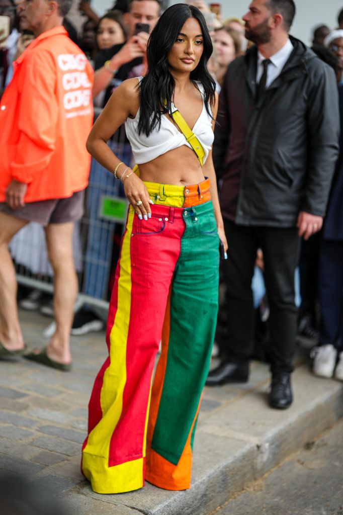 Amina Muaddi wears a white V-neck / rolled-up waist / cropped tank-top, a yellow shiny leather Puzzle crossbody bag from Loewe, green / red / yellow / orange colored denim wide legs jeans pants with trompe l'oeil waist, pumps heels shoes, gold bracelets, outside the Loewe show, during Paris Fashion Week - Menswear Spring/Summer 2023,