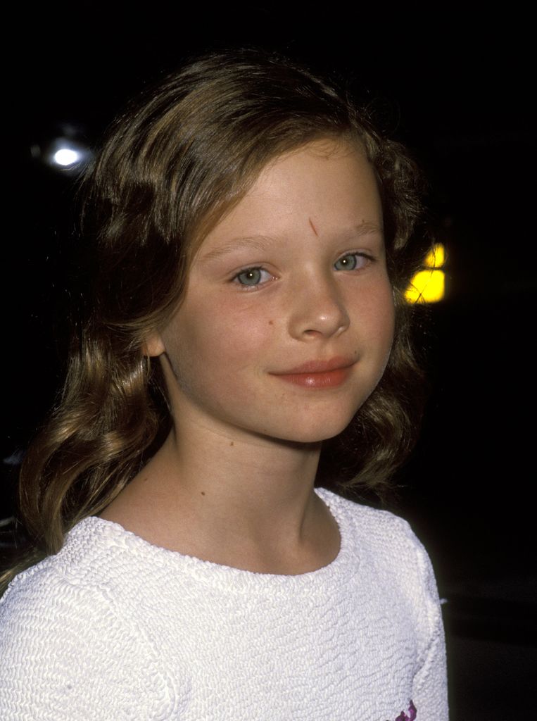 Thora Birch as a child in a white top smiling for the camera shyly