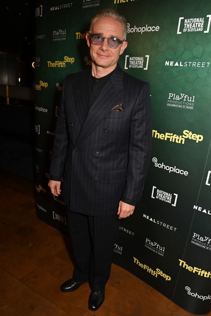 Martin Freeman in a dark suit and glasses in front of green step-and-repeat photographer background while posing for cameras at industry event