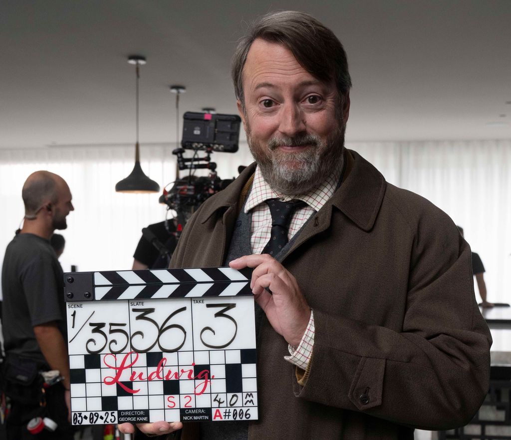 actor in shirt and tie and coat holding clapperboard on TV set