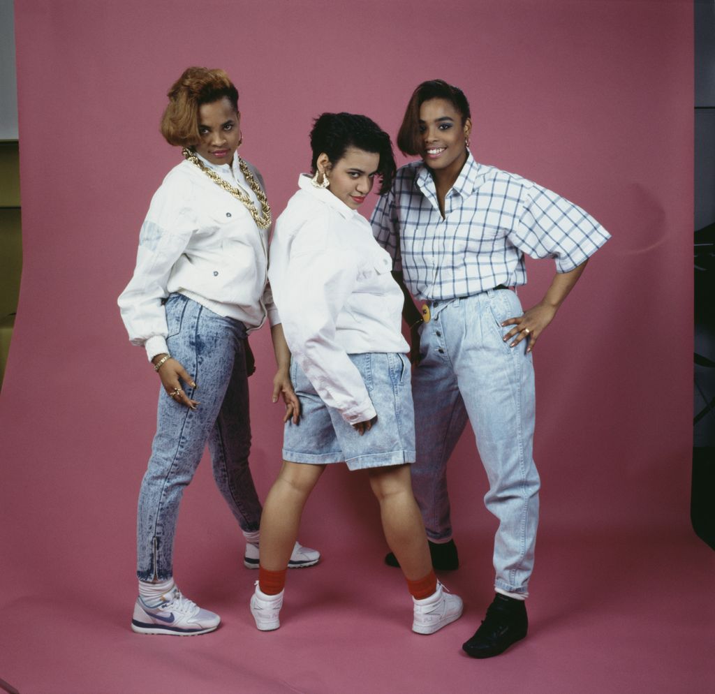 Three stylish women in iconic 1980s hip-hop fashion, wearing acid-wash denim, oversized white jackets, gold jewelry, and high-top sneakers, posing confidently against a pink studio backdrop.
