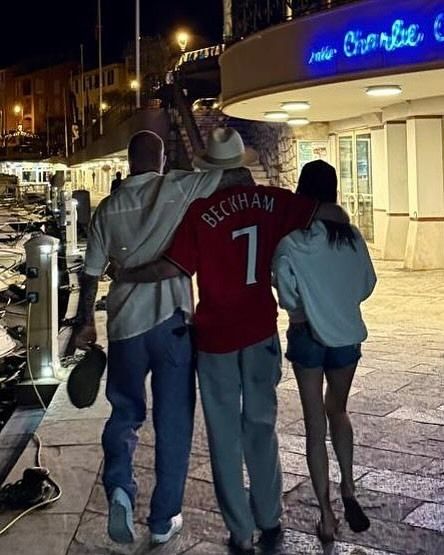 VB walking at night by sea in denim shorts alongside her husband and son