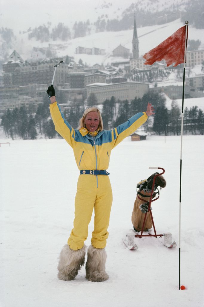 A woman in a bright yellow and blue ski suit with furry snow boots poses cheerfully on a snowy mountain backdrop, representing bold 1980s ski fashion and winter luxury style.