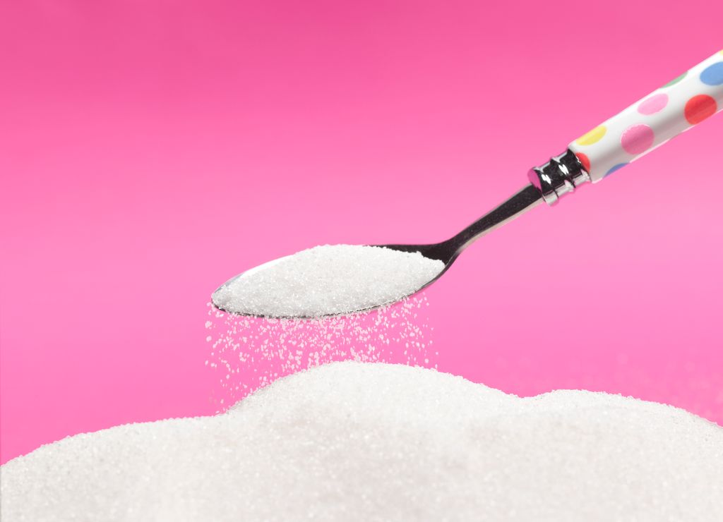 A spoon shaking sugar onto another pile of sugar on a pink background