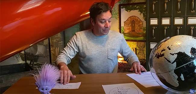 jimmy sits at an office desk looking at piles of paper with a bright orange indoor tunnel slide in the background and a lilac hued troll doll on his desk