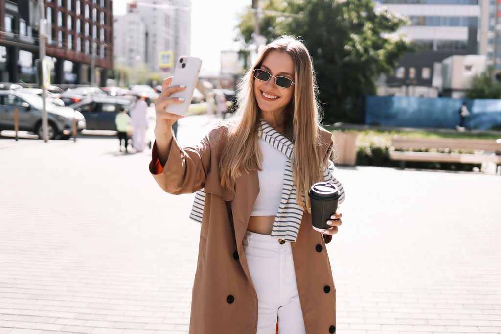 Portrait of a smiling young blonde woman in sunglasses and fashionable clothes walking through the streets of the city holding a glass drinking hot coffee outdoors. A beautiful millennial student girl communicates using a mobile phone and wireless technology takes selfies and laughs while looking at the camera