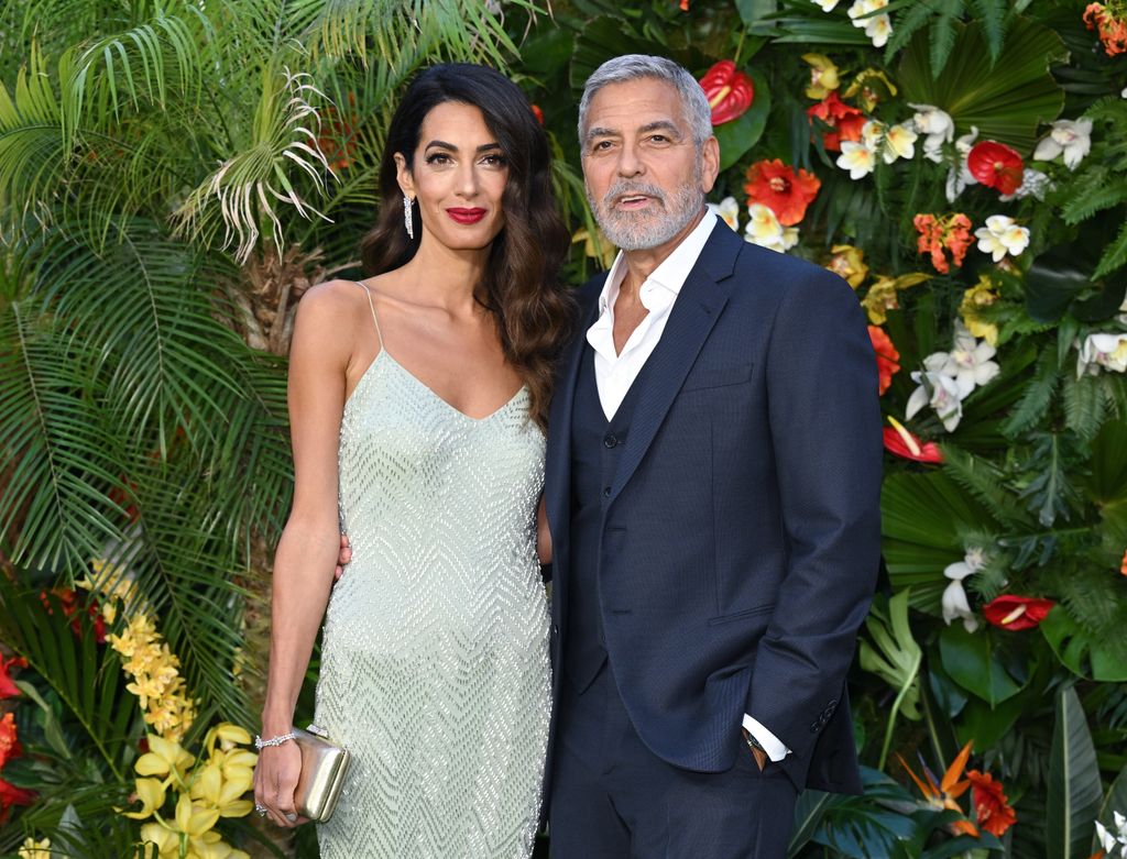 George Clooney dan Amal Clooney berdiri bersama di depan tanaman dan latar belakang pohon saat menghadiri pemutaran film film