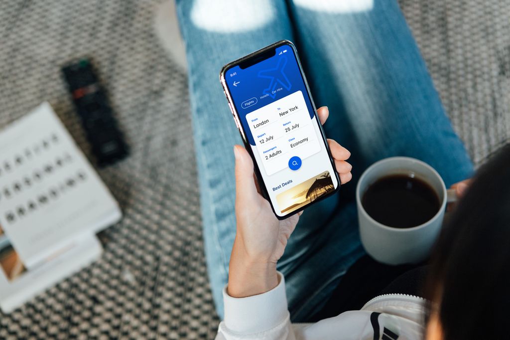 Over the shoulder view of a young woman using smartphone to book flight tickets and plan holiday. She is holding a coffee