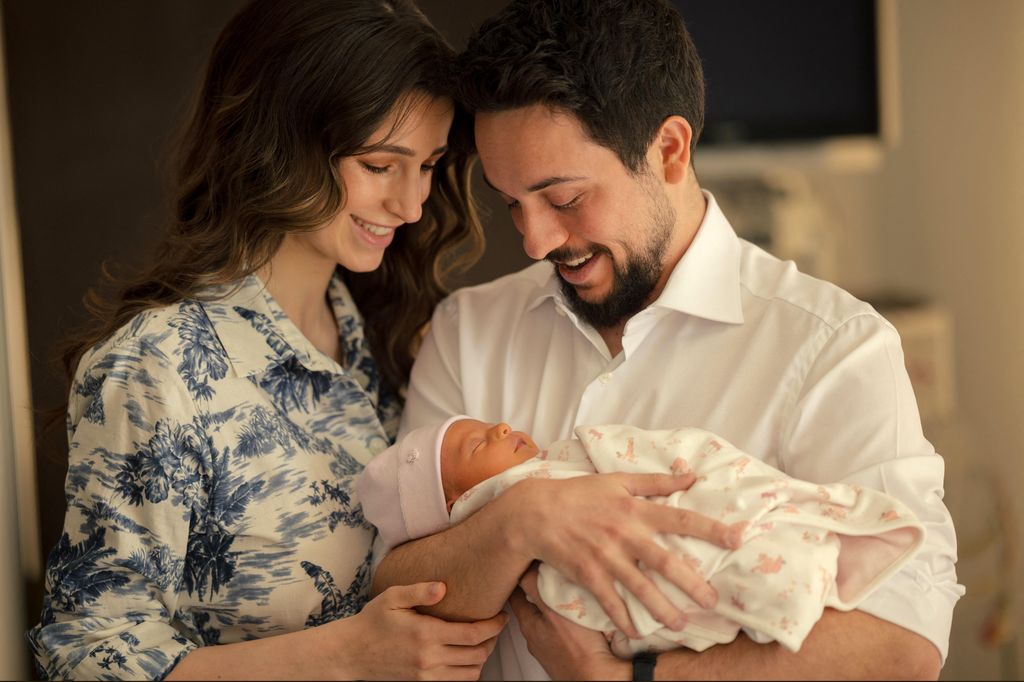 Princess Rajwa in blue and white printed dress beside prince hussein holding baby Princess Iman in blanket