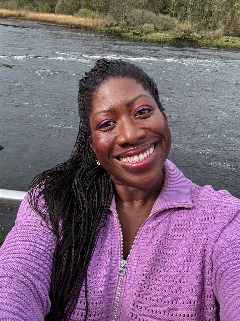 Woman posing and smiling in a purple zip up jumper in front of a lake
