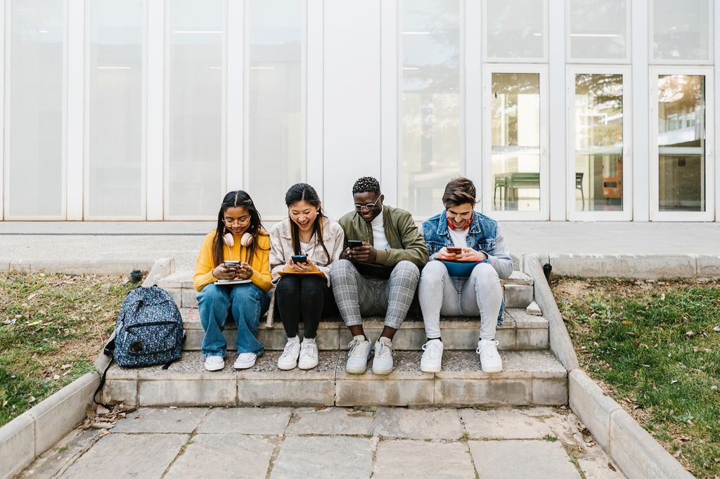 Young multiracial students using cell phone sitting at campus school 