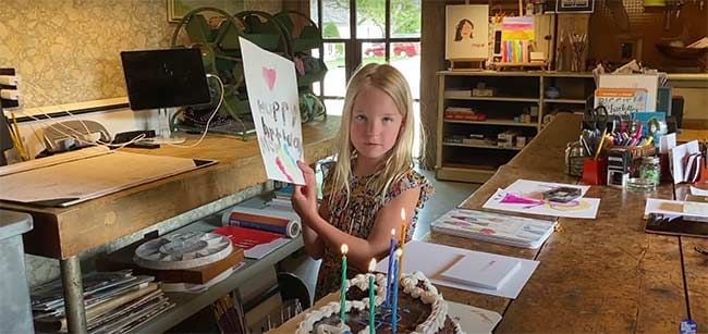 a child holds up an abstract looking painting that they appear to have created themselves while standing in a huge art workshop with paint pots and brushes everywhere 