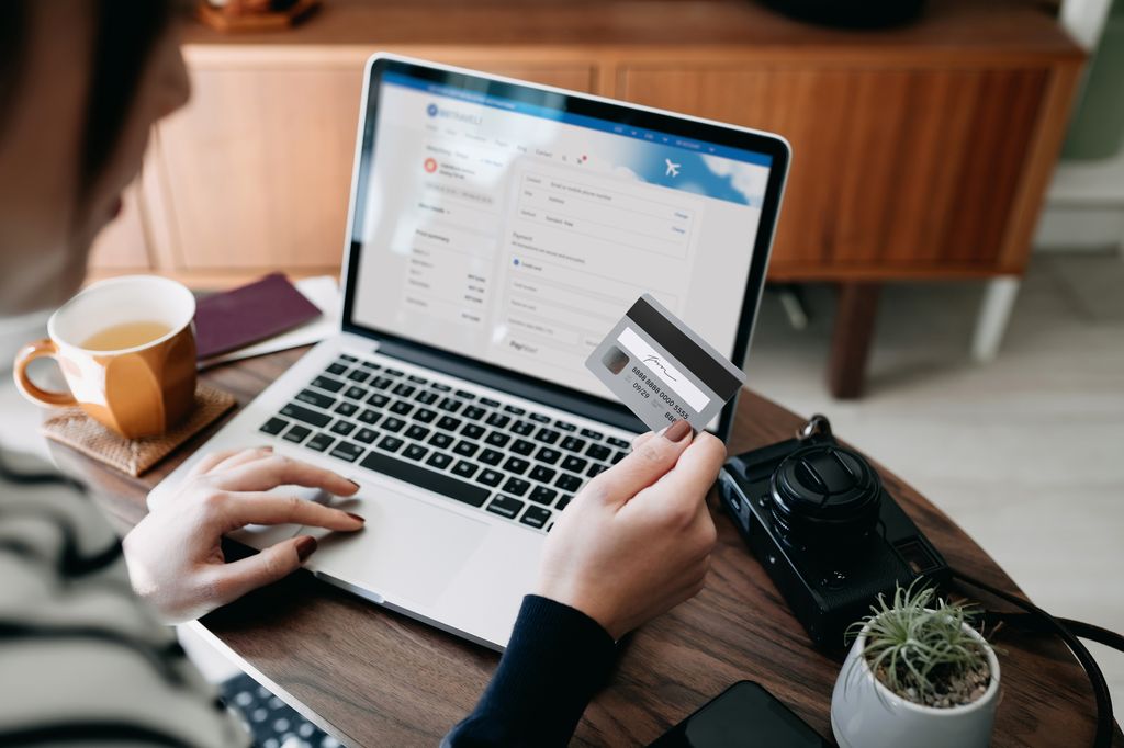 Over the shoulder view of young Asian woman shopping online for flight tickets on airline website with laptop, entering credit card details to make mobile payment at home 