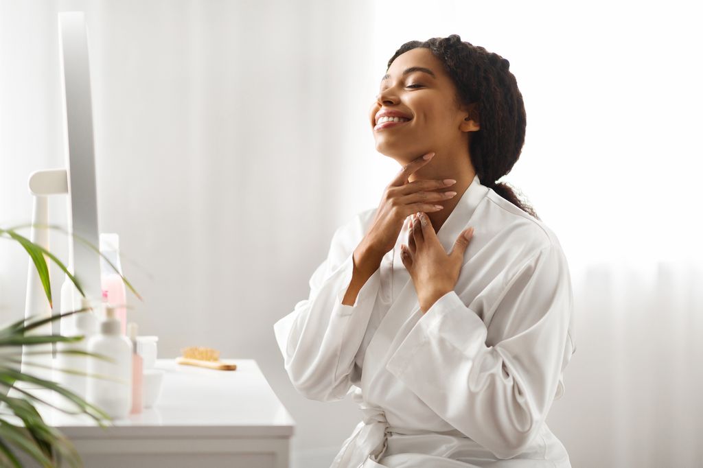 Beautiful Black Woman Touching Her Neck, Making Skin Lifting Massage At Home, Smiling Happy Young African American Female Wearing White Silk Robe Sitting At Dressing Table In Bedroom, Copy Space
