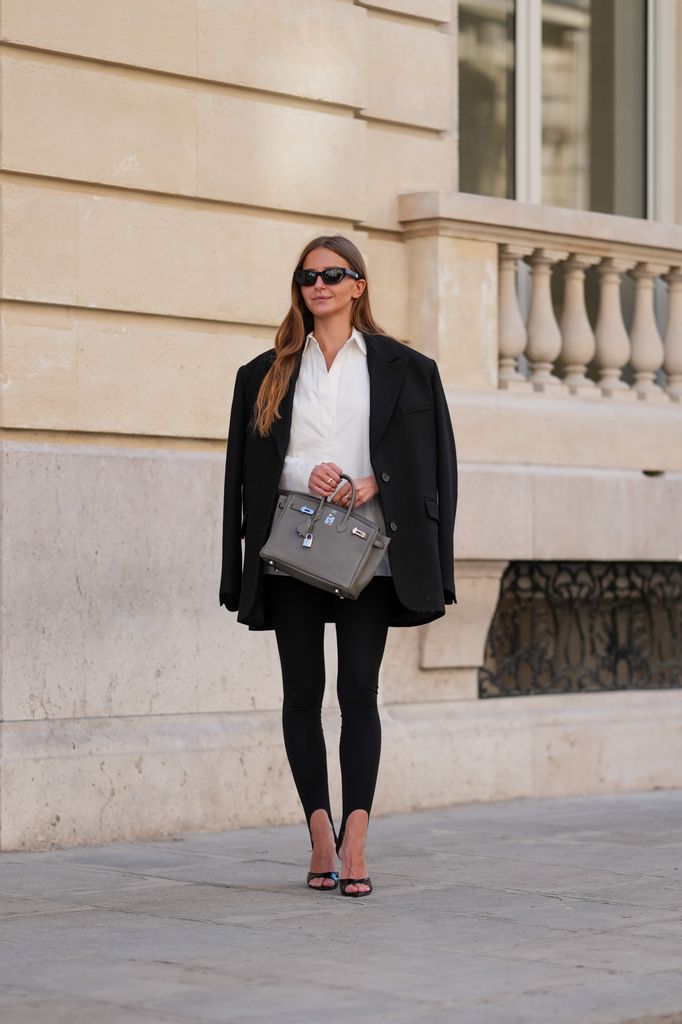 Diane Batoukina mengenakan kacamata hitam Oliver Peoples, rambut panjang lurus berwarna coklat dengan belahan tengah, blus/kemeja Autentiments putih dengan kerah terbuka, blazer Autentiments hitam yang disampirkan di bahu, dan celana/legging Autentiments hitam. Tas tangan Hermes birkin berbahan kulit abu-abu dibawa dengan tangan, dipadukan dengan sepatu hak Tony Blanko ujung terbuka dari kulit hitam dengan tumit ramping. Bidikan seluruh tubuh, saat sesi foto fesyen gaya jalanan, pada 28 April 2025 di Paris, Prancis. (Foto oleh Edward Berthelot/Getty Images)