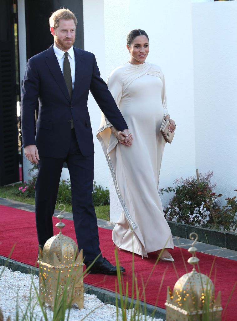 Prince Harry in suit walking with Meghan in ivory caped dress