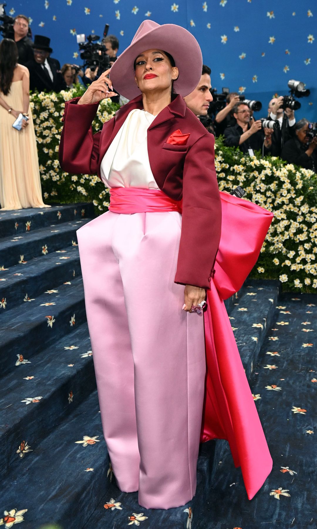 Tracee Ellis Ross wears oversize blush pink satin trousers with fuschia bow detail at the back, a cropped deep red jacket and dusky pink hat