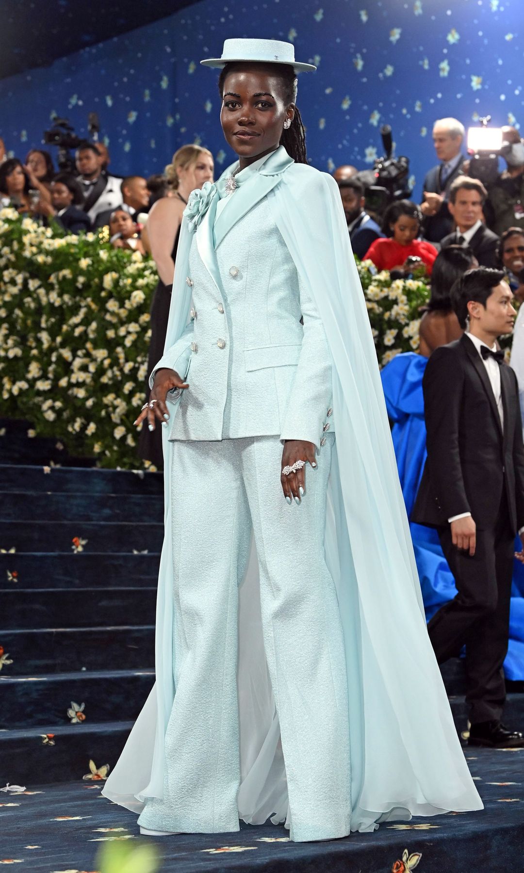 Lupita Nyong'o wears an icy blue suit with double breasted blazer and matching hat and corsage.